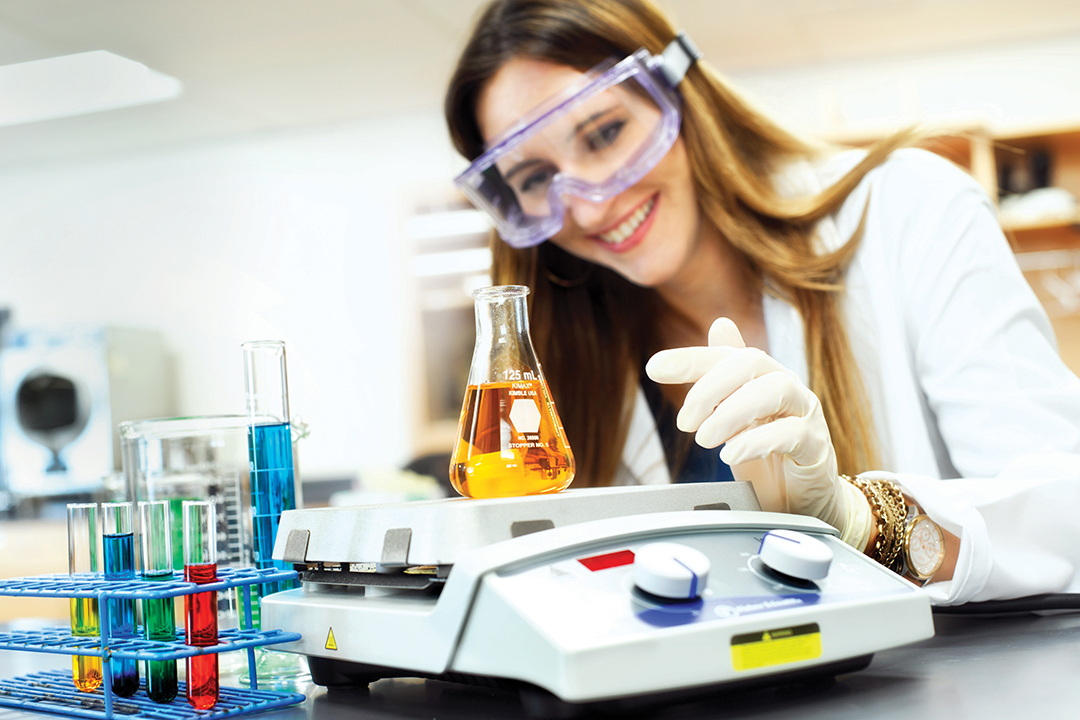 student in a lab with goggles looking at an orange liquid in a beaker on a scale
