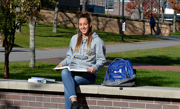 student sitting on a brick wall with a book wearing a SUNY Poly sweatshirt